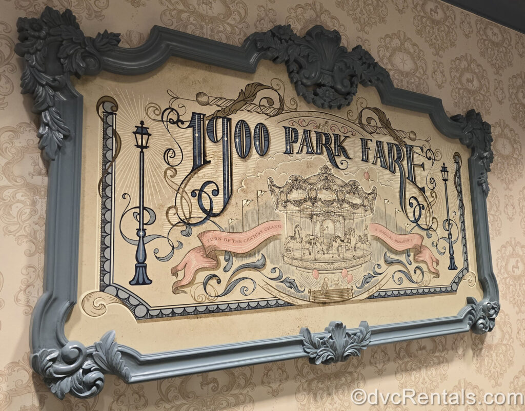 The sign for 1900 Park Fare at the Villas at Disney’s Grand Floridian Resort & Spa. The sign has a Victorian design with 1900 Park Fare written in a blue font, and a black and white carousel drawing in the center. On either side of the carousel there are old-fashioned streetlamps, and a pink banner reading “turn of the century charm. A magical menagerie.” This sign is framed by an ornate blue frame with beige damask wallpaper behind it.