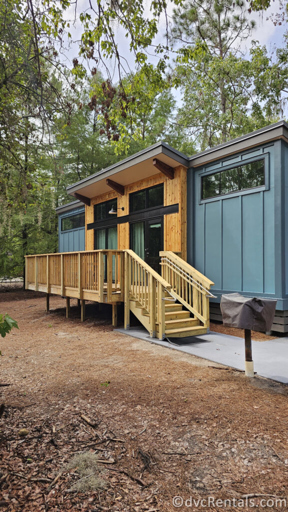 A blue cabin with a wooden deck and stairs. The cabin has large windows and a modern design. There is a grill on the ground below the deck, and the cabin is surrounded by trees.