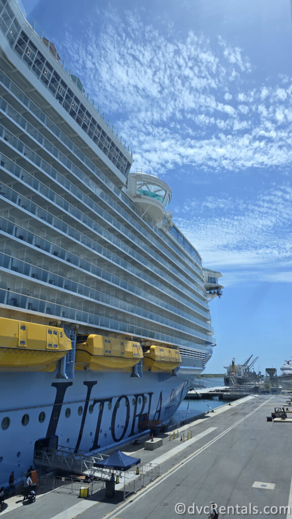 The white exterior of Royal Caribbean’s Utopia of the Seas ship docked in Fort Lauderdale under a blue sky. “Utopia” is written in large navy-blue letters along the side of the ship with yellow lifeboats visible above.