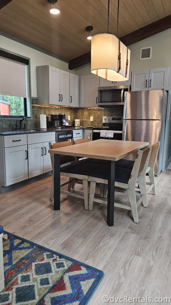 A kitchen and dining area in a cabin. The kitchen features white cabinets, stainless steel appliances, and a wooden countertop. A large dining table with chairs is located in the center of the room.