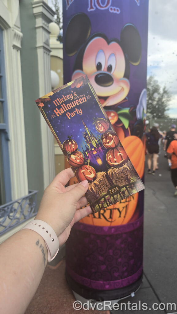 A person’s hand is holding a Mickey’s Not-So-Scary Halloween Party 2024 map that is decorated with 6 jack-o-lanterns and Cinderella Castle in shares of purple and green. There is also a large purple sign with Mickey Mouse in the background.