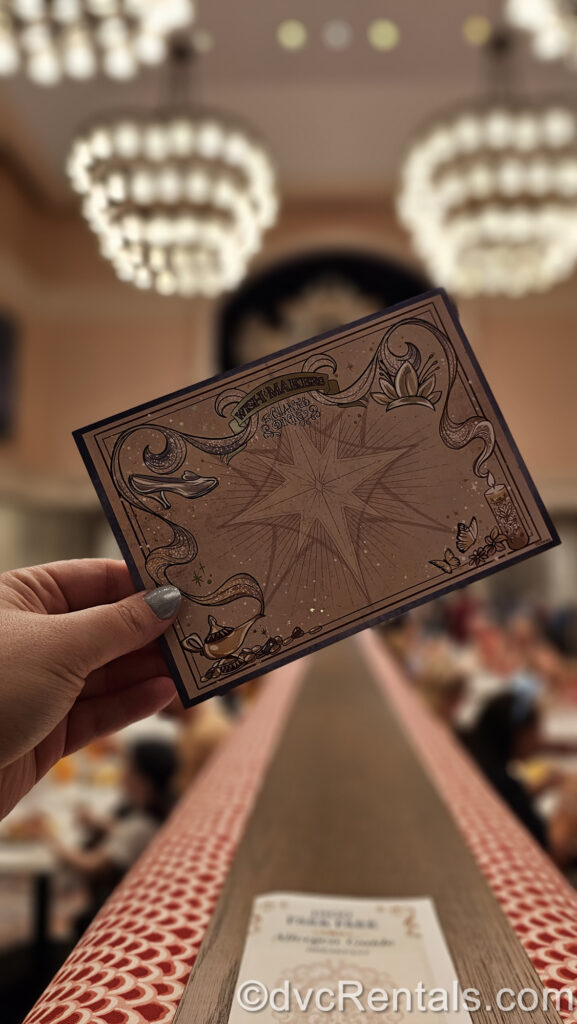 A hand holds a Wish Maker card at 1900 Park Fare. The card is beige with a large star symbol in the center. Around the edge of the card there is a blue ribbon design connecting Cinderella’s glass slipper, Tiana’s tiara, the candle from Encanto, and the Genie’s lamp from Aladdin.