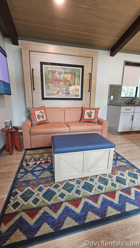 A living room area in a cabin with a wood-paneled ceiling and a large, folding bed with a vintage poster above it. There is a couch with pillows, a coffee table, and a colorful rug on the floor.