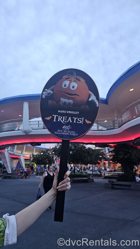 A Magic Kingdom Cast Member is holding a sign in Tomorrowland indicating a trick-or-treat trail location. The sign is black and navy-blue and features an orange M&M candy character made to look like a pumpkin. The sign reads “Mars Wrigley Treats! Mickey’s Not So Scary Halloween Party!” in white lettering