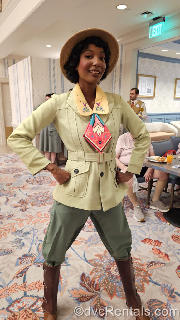 Princess Tiana stands smiling with her hands on her hips at 1900 Park Fare. Tiana is wearing a soft green jacket with a yellow collar, red and blue kerchief, a light brown hat, olive green pants, and brown knee-high boots