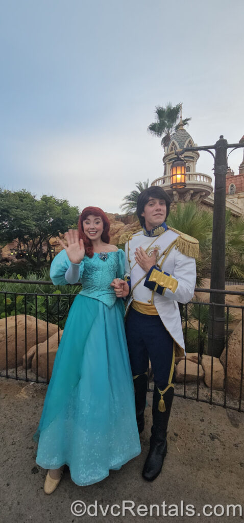 Princess Ariel and Prince Eric meet and greet in Fantasyland. Ariel is wearing a teal ballgown and waving to the camera. Eric is wearing a white and blue military-dress style suit with black riding boots.