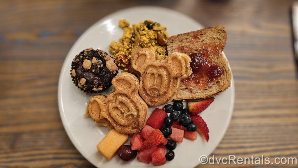 A selection of breakfast foods from 1900 Park Fare on a white plate. On the plate is a chocolate muffin, muesli, brown bread toast with strawberry jam, fresh fruit, and 2 Mickey waffles