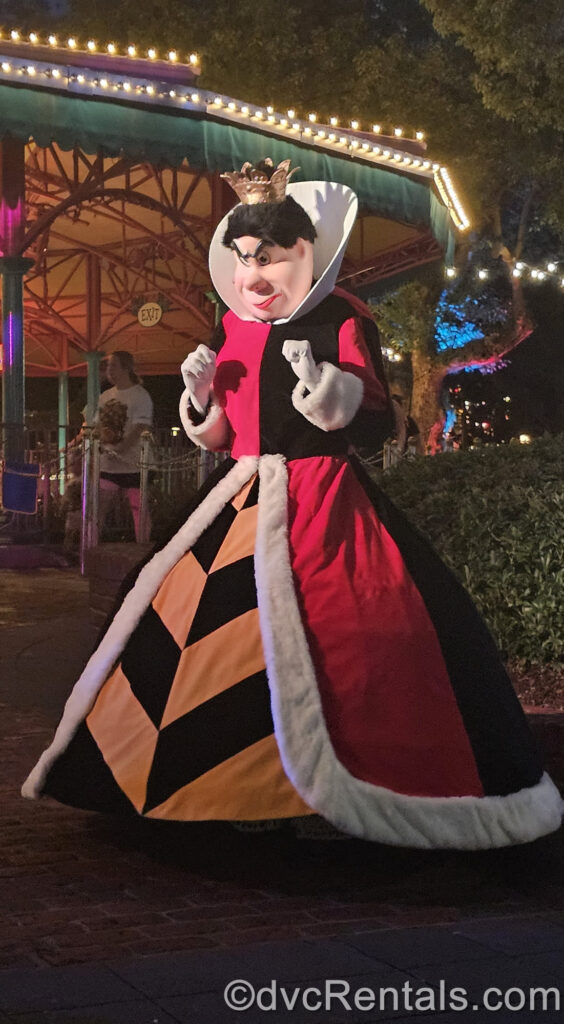 The Queen of Hearts in front of Mad Tea Party in Fantasyland. She wears a small gold crown on her head, white gloves on her hands, and her dress is red and black with gold and black stripes and white accents around the skirt.