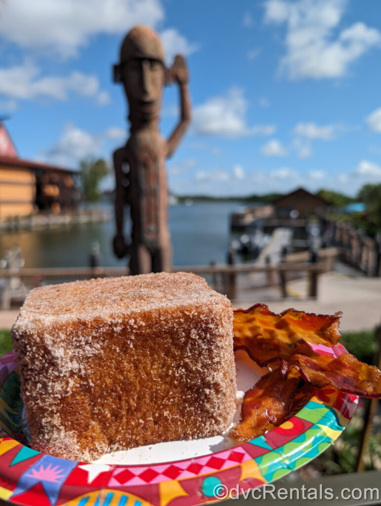 An order of thick, golden brown Tonga Toast and bacon on a colorful plate from Capt. Cook’s shown in the outside seating area. Behind the plate of food there is a tiki-style statue and the boat dock at Disney’s Polynesian Villas & Bungalows.