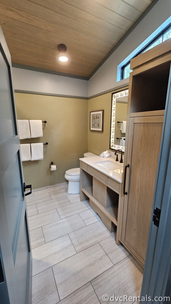 A bathroom with a white toilet, a wooden vanity with a white countertop, and a large mirror. The walls are painted yellow with a wood accent, and the ceiling is wood-paneled.