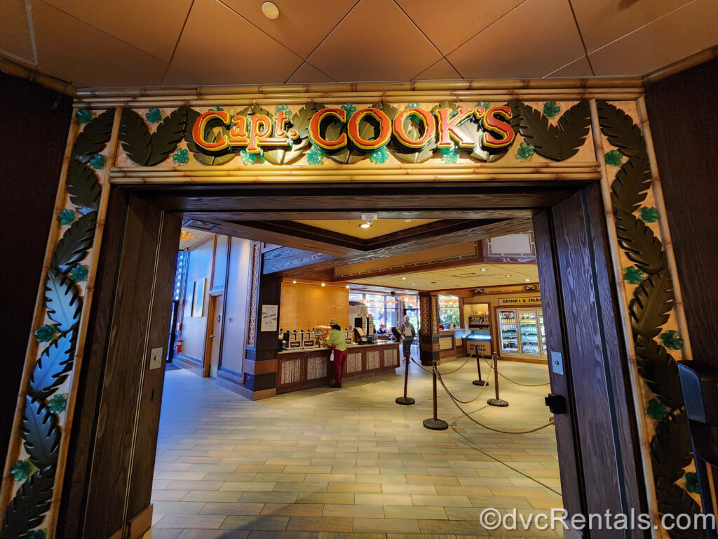 The interior entry way leading into Capt. Cook’s. The outside of the door frame is decorated with green palm leaves and flowers, and the restaurant’s name is shown at the top in orange writing.