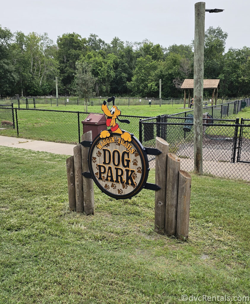 A circular sign made of metal and wood with the words "BARKING TRAILS DOG PARK" written on it. The sign is surrounded by a wooden fence and is located in a grassy area with trees in the background.