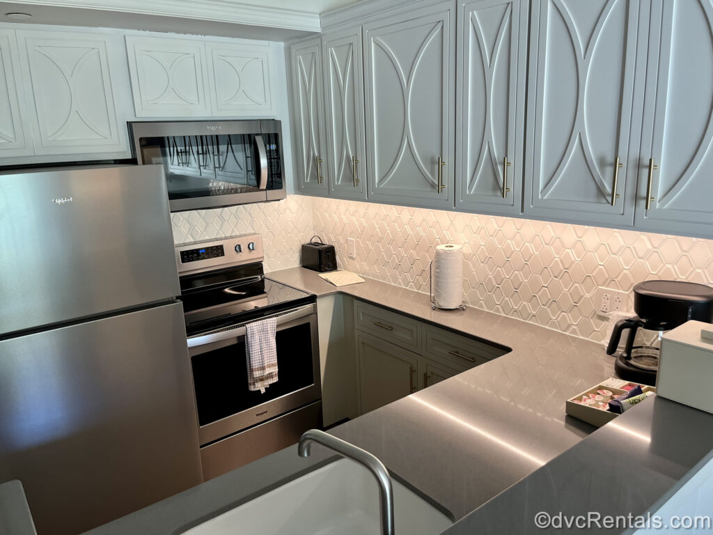 The kitchen in a Two Bedroom Villa at Disney’s Saratoga Springs Resort & Spa. The cabinets are white with a geometric pattern, and the backsplash is also white geometric tiles. The counter is dark grey with a white sink. The refrigerator, microwave, and stove are all stainless-steel. There is a black toaster, roll of paper towels, a black coffee maker, and cream and sugar on the counter.