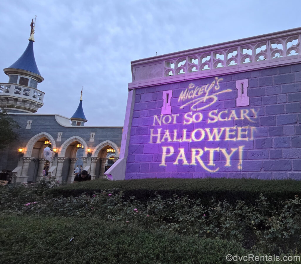 The words “Mickey’s Not So Scary Halloween Party!” are projected in white light onto the grey stone bricks of a building façade in Fantasyland at Magic Kingdom.