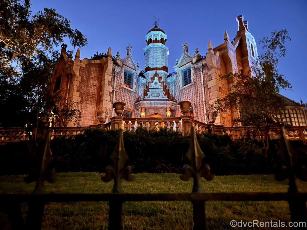 Haunted Mansion at Magic Kingdom in the evening. The red brick façade of the mansion is illuminated blue and orange, and the black wrought iron spikes of the fence in front of the attraction are visible in the foreground.