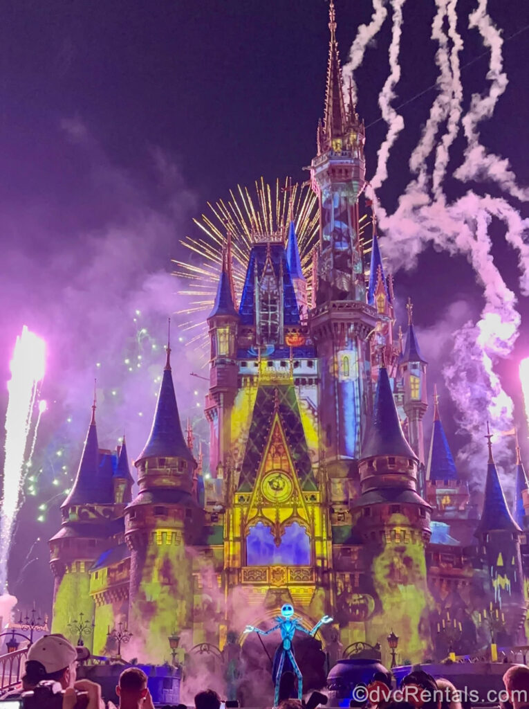 A large black and white Jack Skellington puppet performs in front of Cinderella Castle at night. The castle is lit up with yellow projection effects and golden fireworks pop behind it.