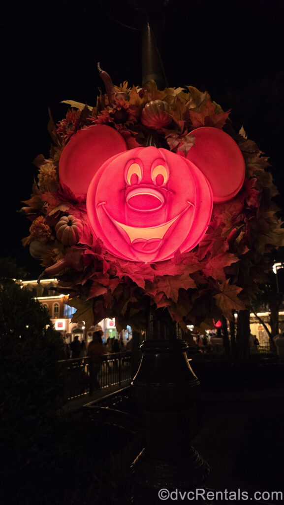 A bright orange Mickey-shaped pumpkin light glows at Magic Kingdom at night. The cheerful face carved on the pumpkin is illuminated and a wreath of falls leaves and gourds surrounds it.