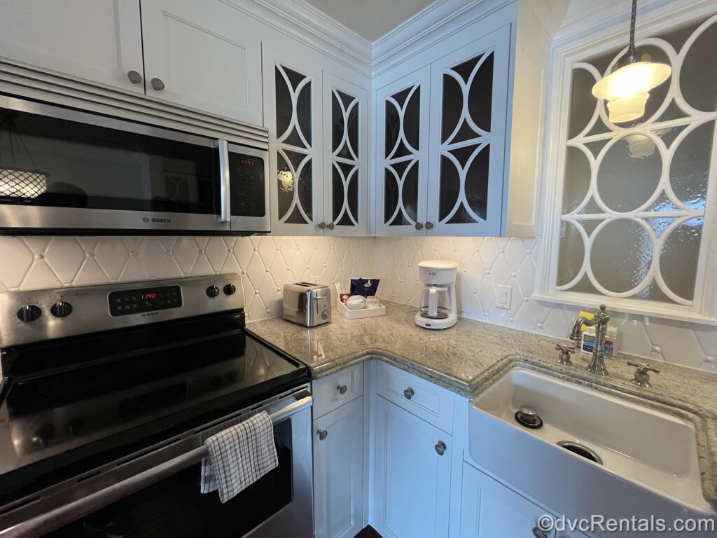 A full kitchen at the Villas at Disney’s Grand Floridian Resort & Spa. The upper and lower cabinets are white with a circular cut-our pattern on the upper cabinets. The backsplash is also white with diamond-shaped tiles. The counter is a light speckled grey with a white sink. The microwave and stove are both stainless-steel. There is a silver toaster, a white coffee maker, and coffee packets, cream, and sugar on the counter.