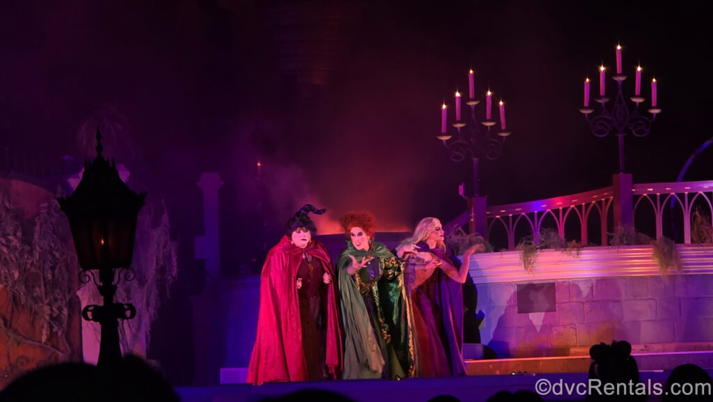 The Sanderson Sisters perform on stage. Mary has dark hair twirled upward and is wearing a red cloak, Winifred has red curly hair and a green cloak, and Sarah has long blonde hair and a purple cloak. There is purple and pink lighting 2 candelabras glow in the background.