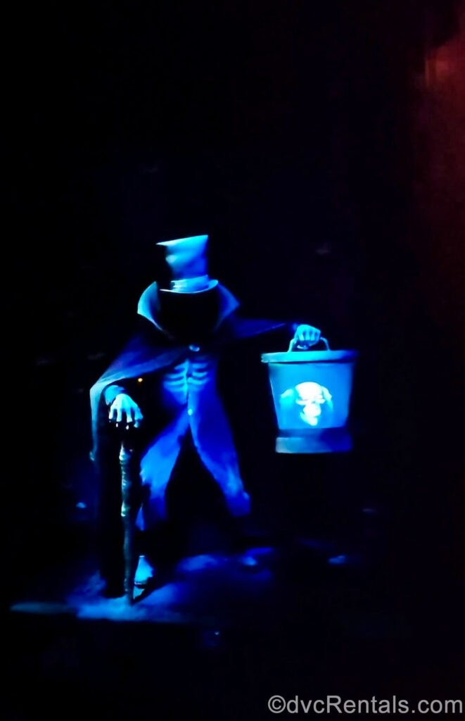 The shadowy, faceless figure of the Hatbox Ghost appears to be holding a carrying case in which you can see his spooky severed head. The Hatbox Ghost wears a suit, cape, and top hat and also holds a cane, and is lit up blue in the darkness of the attraction setting.