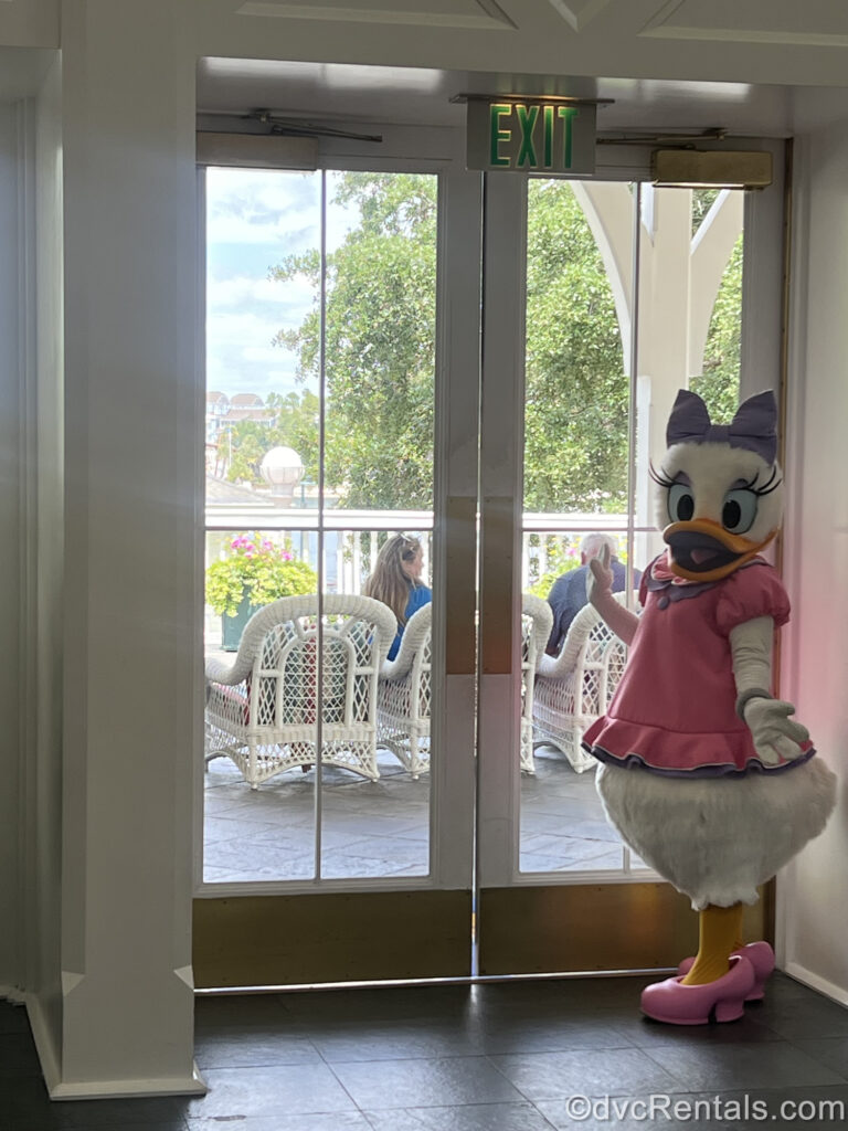 Daisy Duck stands at glass double doors leading out to a seating area at Disney’s BoardWalk Villas. Daisy is wearing a pink shirt and shoes, with a purple bow. Outside the doors, guests can be seen sitting in white wicker armchairs.