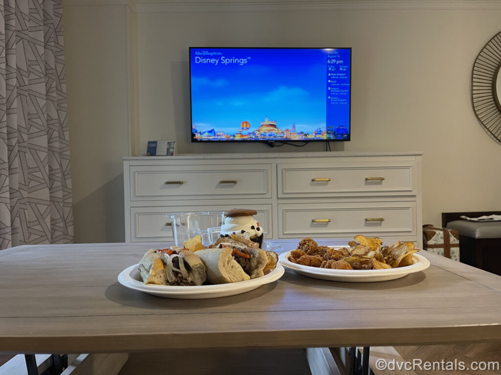 2 paper plates of food on the coffee table in a Studio at Disney’s Saratoga Springs Resort & Spa. The plate on the left has sandwiches and kettle chips on it, with a cupcake with white icing behind it. The plate on the right has chicken nuggets and kettle chips on it. In the background the TV is on the blue resort channel atop the white dresser.