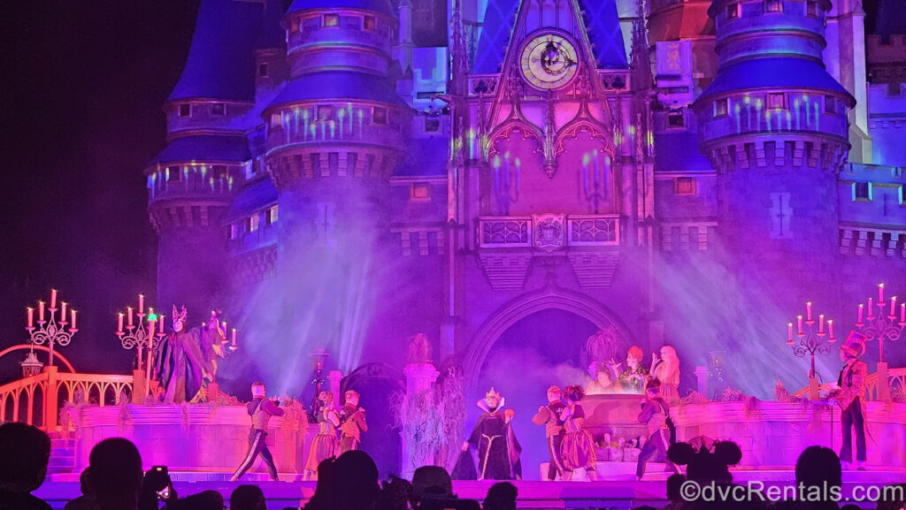 Maleficent, the Evil Queen, the Sanderson Sisters, and Dr. Facilier perform on stage with an ensemble of dancers in front of Cinderella Castle. The castle has spooky candle projections on it and fog rolls over the stage which is illuminated with pink and purple lighting effects.