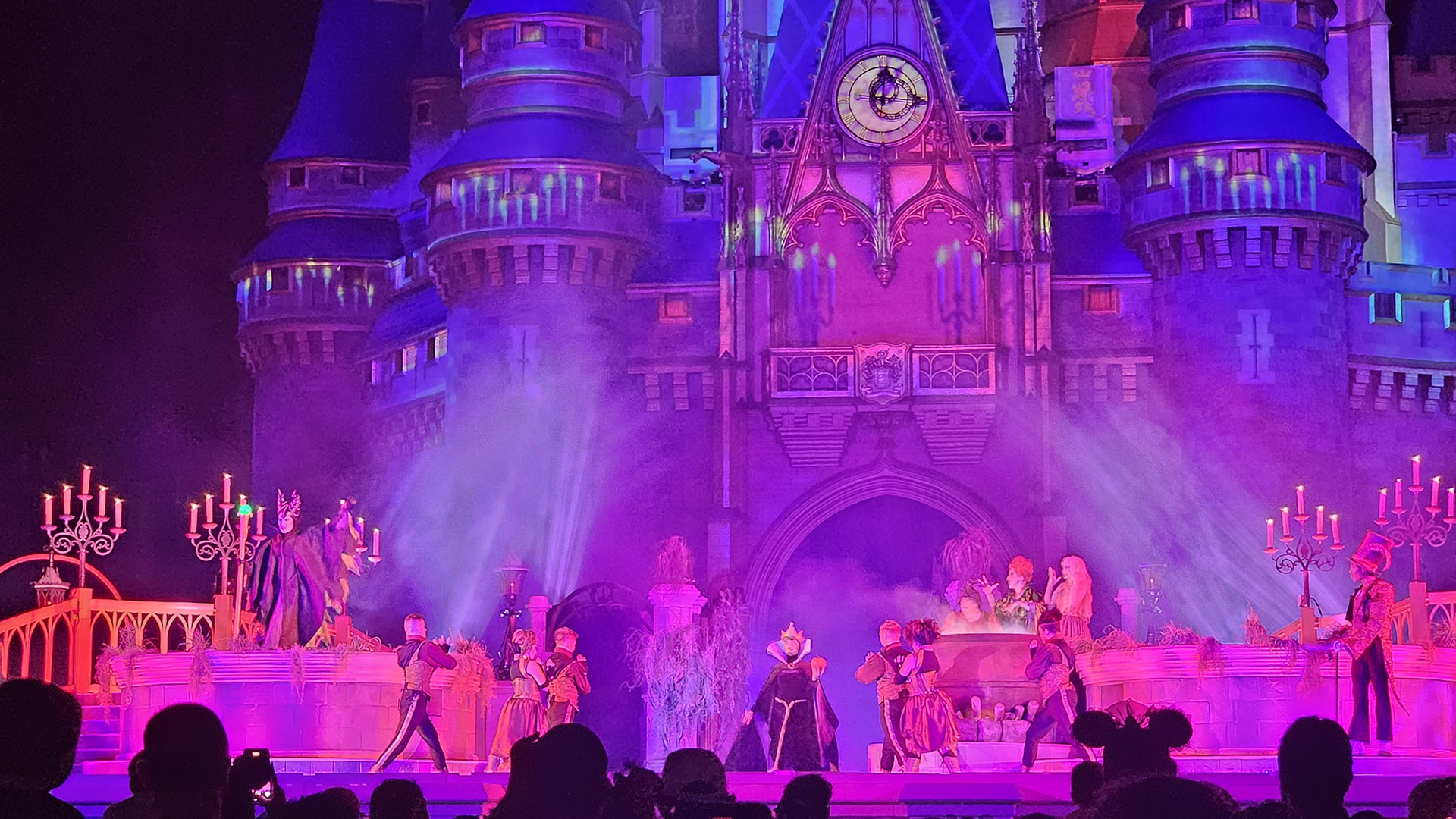 Maleficent, the Evil Queen, the Sanderson Sisters, and Dr. Facilier perform on stage with an ensemble of dancers in front of Cinderella Castle. The castle has spooky candle projections on it and fog rolls over the stage which is illuminated with pink and purple lighting effects.