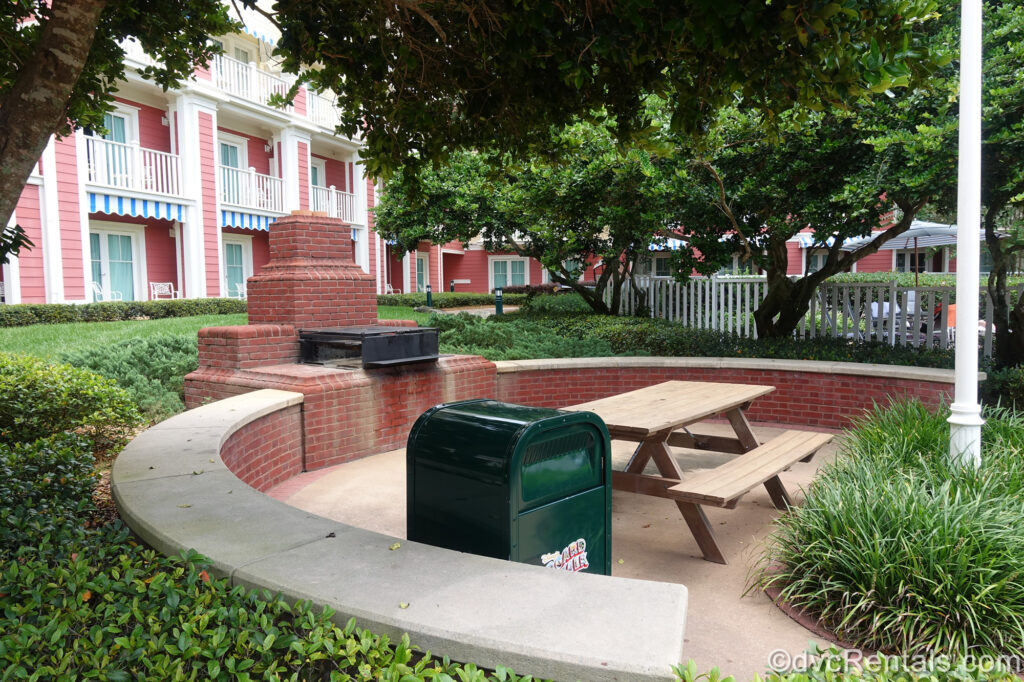 A grilling area at Disney’s BoardWalk Villas. Green foliage surrounds the grill area which is made up of red bricks and a concrete half wall. The grill sits atop the red bricks, and a wooden picnic table is across from it. There is also a green trash can nearby, and the salmon-colored resort building is visible in the background.