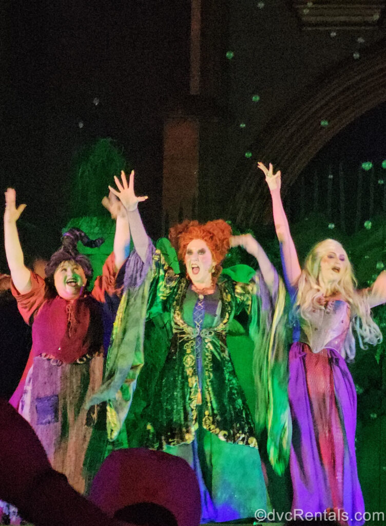 The Sanderson Sisters are performing on stage. Mary has dark hair twirled upward and is wearing an orange and red dress, Winifred has red curly hair and a green cloak, and Sarah has long blonde hair and a purple cloak. There is green lighting in the background and all 3 characters have their arms raised.