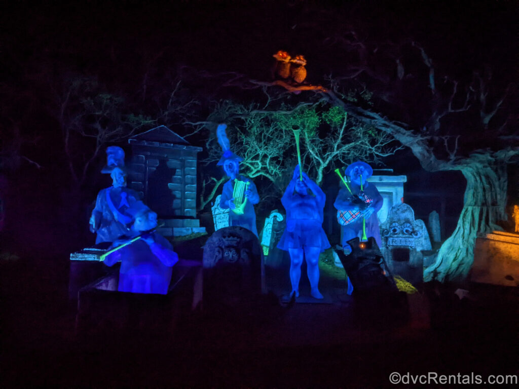 Five semi-transparent, blue-tinted ghost figures play various instruments in a band in the dark Haunted Mansion graveyard. There are grey tombstones surrounding them, and 2 owls with glowing red eyes sit in the tree above them.