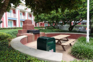 A grilling area at Disney’s BoardWalk Villas. Green foliage surrounds the grill area which is made up of red bricks and a concrete half wall. The grill sits atop the red bricks, and a wooden picnic table is across from it. There is also a green trash can nearby, and the salmon-colored resort building is visible in the background.