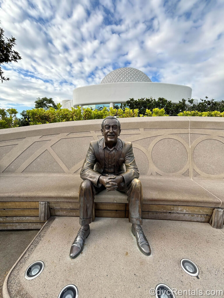 Bronze Walt Disney statue under a blue sky at Dreamers Point in Epcot. The statue is seated on a beige stone bench with part of the Epcot ball icon visible past green foliage and white building facades in the background.