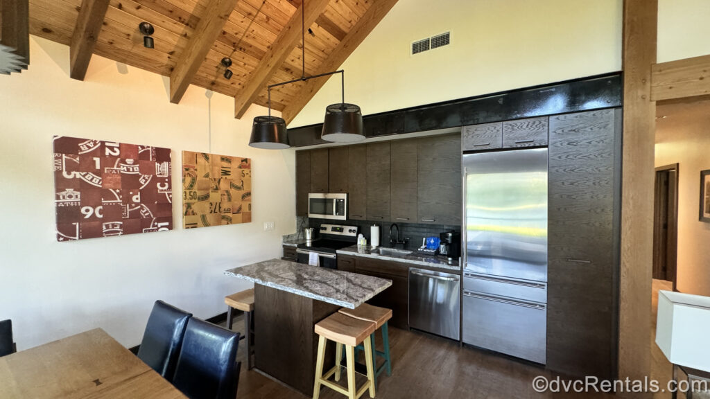 The kitchen of a Cascade Cabin at the Copper Creek Villas & Cabins at Disney’s Wilderness Lodge. The cabinets are dark wood, with a stainless-steel fridge/freezer, dishwasher, and stove. Above the grey stone countertop there is also a stainless-steel microwave. There are paper towels, a silver sink, coffee packets with cream and sugar, and a black coffeemaker on the counter. There is a small table across from it with a grey stone top and wooden stools. There is red and wooden art on the wall, and another wooden table top with black chairs can be seen in the corner.