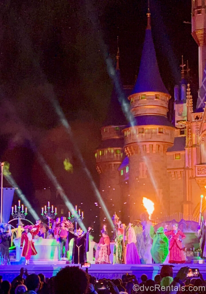 Hades, Captain Hook, Dr. Facilier, the Evil Queen, the Sanderson Sisters, Oogie Boogie and Lady Tremain perform on stage in front of Cinderella Castle, which is lit up orange in the background with special fire effects.