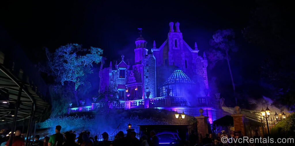 The brick and stone façade of Haunted Mansion is lit up blue and purple at night. In front of the attraction you can see people in queue, and there are glowing streetlamps visible by the ride exit.