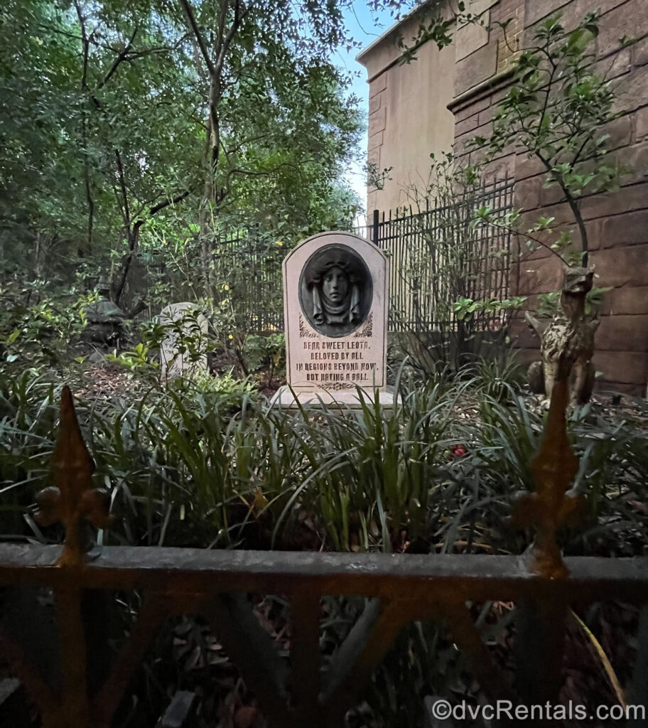 The grey tombstone of Madame Leota from the queue of Haunted Mansion. The tombstone is seen through overgrown green foliage and reads “Dear sweet Leota beloved by all, in regions beyond now but having a ball.” A carving of Madame Leota’s face also decorates the tombstone.