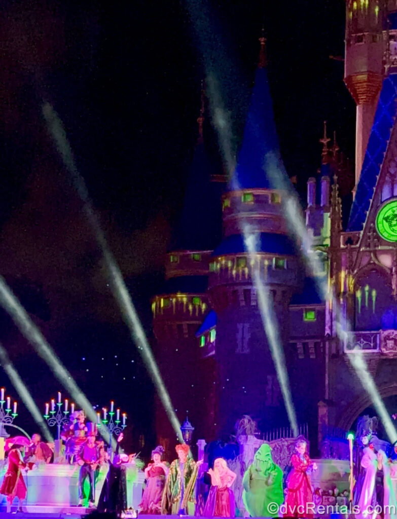 Captain Hook, Dr. Facilier, the Evil Queen, the Sanderson Sisters, Oogie Boogie, Lady Tremaine, Maleficent, and Cruella DeVil perform on stage in front of Cinderella Castle, as white lighting effects shoot upwards.