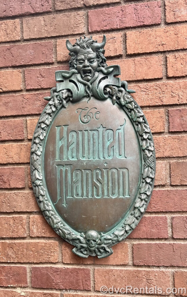 A green and grey metal oval-shaped sign reading “The Haunted Mansion” in a gothic-style font is displayed on a reddish-brown brick wall. At the top of the sign there is a scary face carved with horns, wild hair, and snakes. At the bottom of the sign there is a small skull and crossbones.