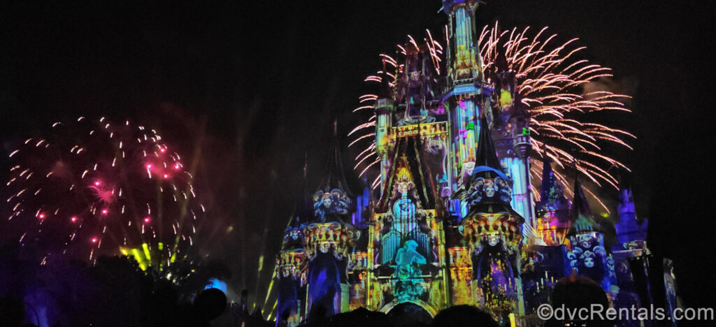 Cinderella Castle is illuminated with spooky and colorful Haunted Mansion-themed projections, as pink and white fireworks burst in the sky.