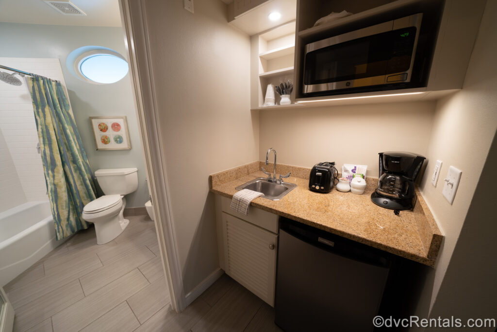 A Studio kitchenette at Disney’s Old Key West Resort. The lower and upper cabinets are white, with a stainless-steel mini fridge at the bottom. The brown countertop has a steel mini sink, black toaster and coffeemaker, cream and sugar and coffee packets on it. Above the counter there is a stainless-steel microwave and disposable cups, utensils, plates, and bowls. To the left of the kitchenette is the doorway into the bathroom.