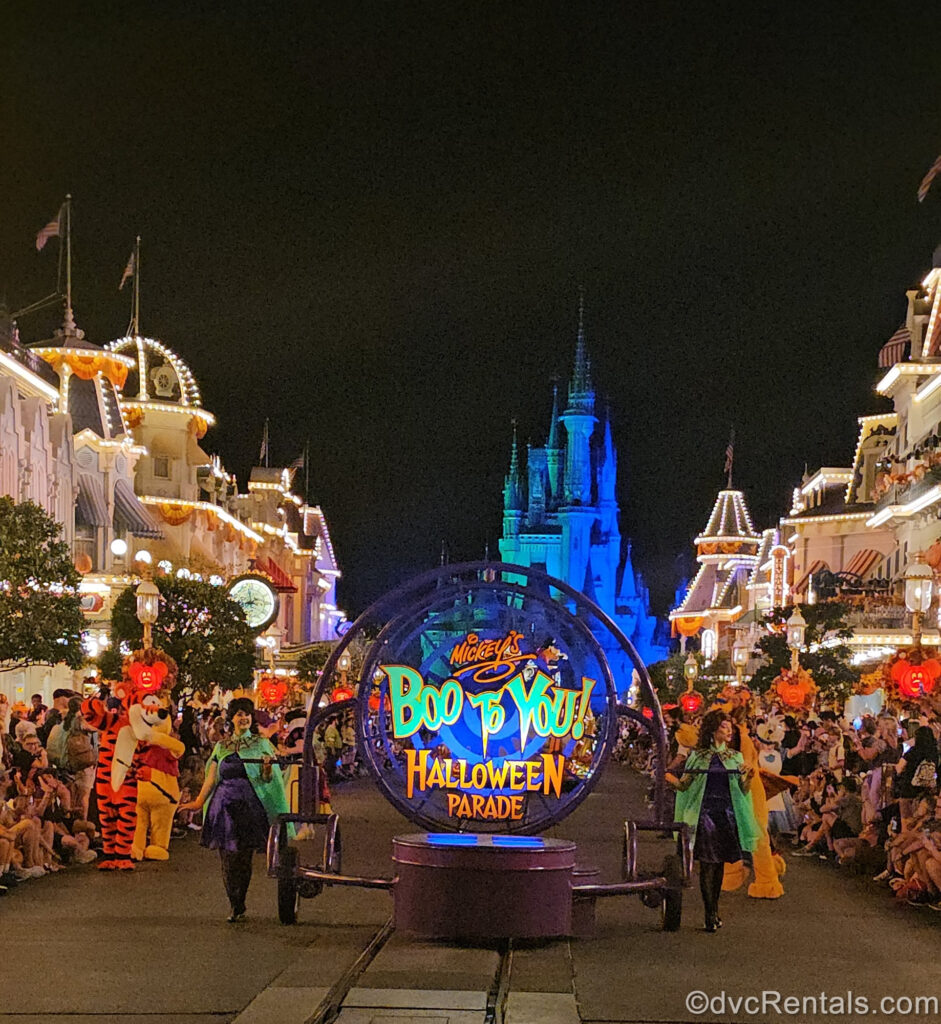 Looking toward Cinderella Castle down Main Street U.S.A. at night with a parade float displaying a sign reading “Mickey’s Boo-to-You! Halloween Parade”. There are Cast Members in green cloaks on either side of the float and crowds on each side of Main Street. Characters Tigger and Winnie the Pooh are seen near the parade float, and Cinderella Castle is illuminated blue in the background.