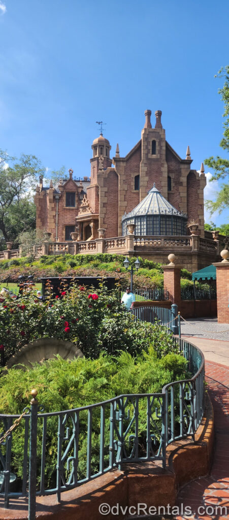 he red brick and beige stone building of the Haunted Mansion is seen in daylight. In front of the mansion there is green foliage made to look overgrown, as well as red roses in the garden that is surrounded by a green-grey wrought iron fence.