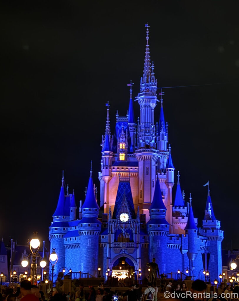 Cinderella Castle at Magic Kingdom at night. Crowds of park guests are seen in front of the castle which is lit up blue with pink accent lighting.