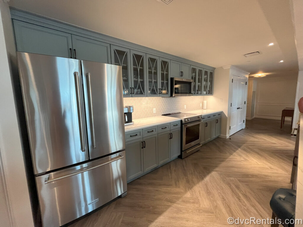 The spacious kitchen in a Grand Villa at Disney’s Riviera Resort. There is a large stainless-steel refrigerator with double doors, slate blue cabinets, a stainless-steel stove and microwave. The center, upper cabinets have cut outs displaying the dishware. The countertop and backsplash are a creamy white color with a coffeemaker and paper towels on the counter.