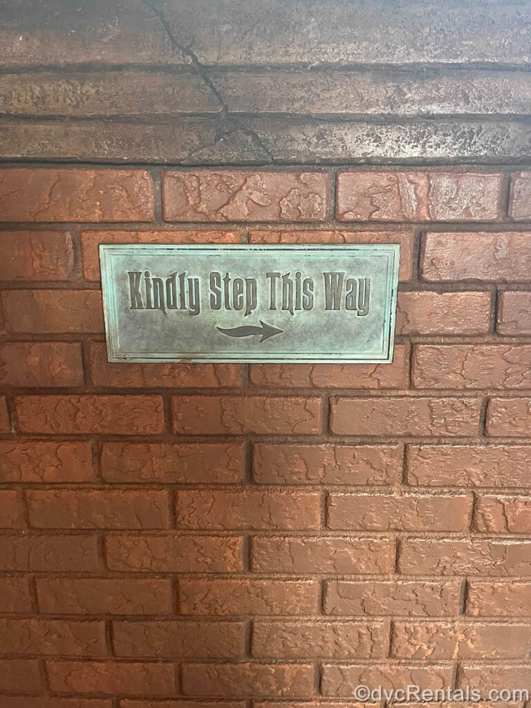 A worn-looking metal sign reading “Kindly Step This Way” in a gothic-style font with a small right-pointing arrow below the wording is posted on a reddish-brown brick wall.