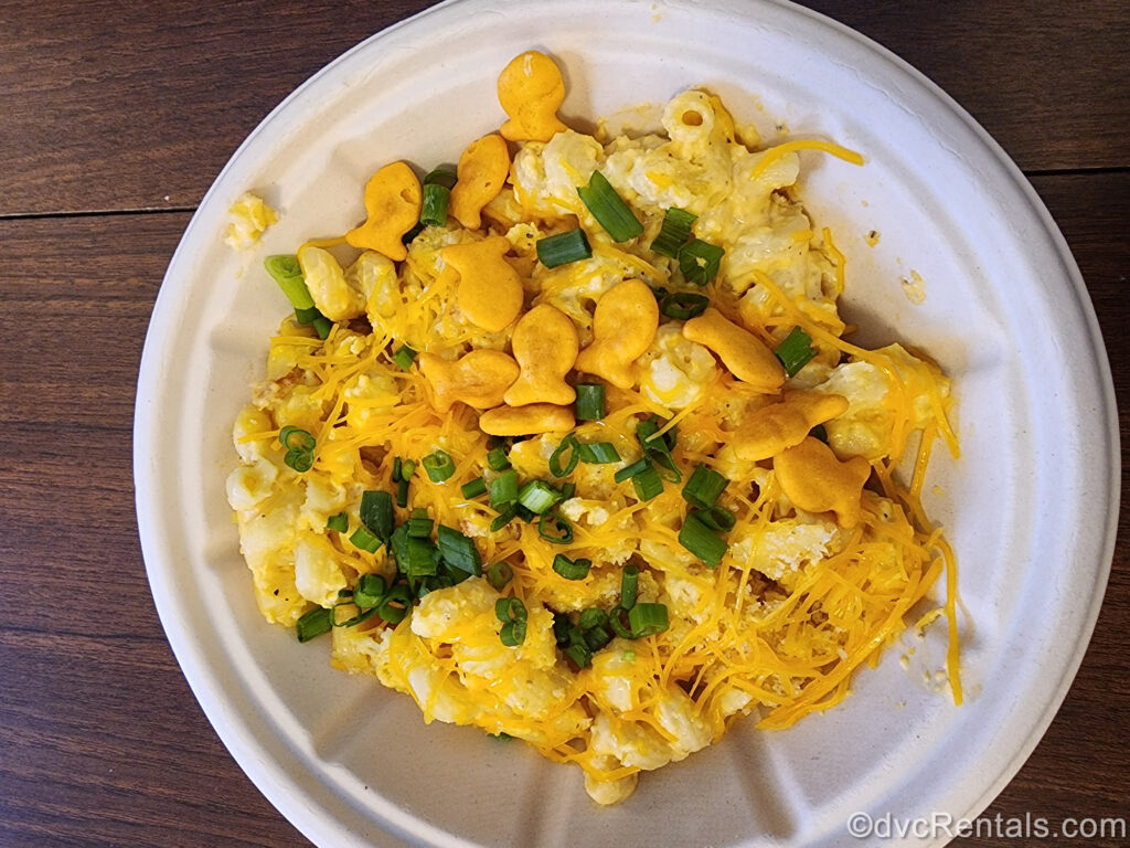 Macaroni and Cheese from the Beach Club Marketplace served in a paper to-go bowl. The pasta is topped with shredded cheddar cheese, small green onion pieces, and cheese-flavored Goldfish crackers.