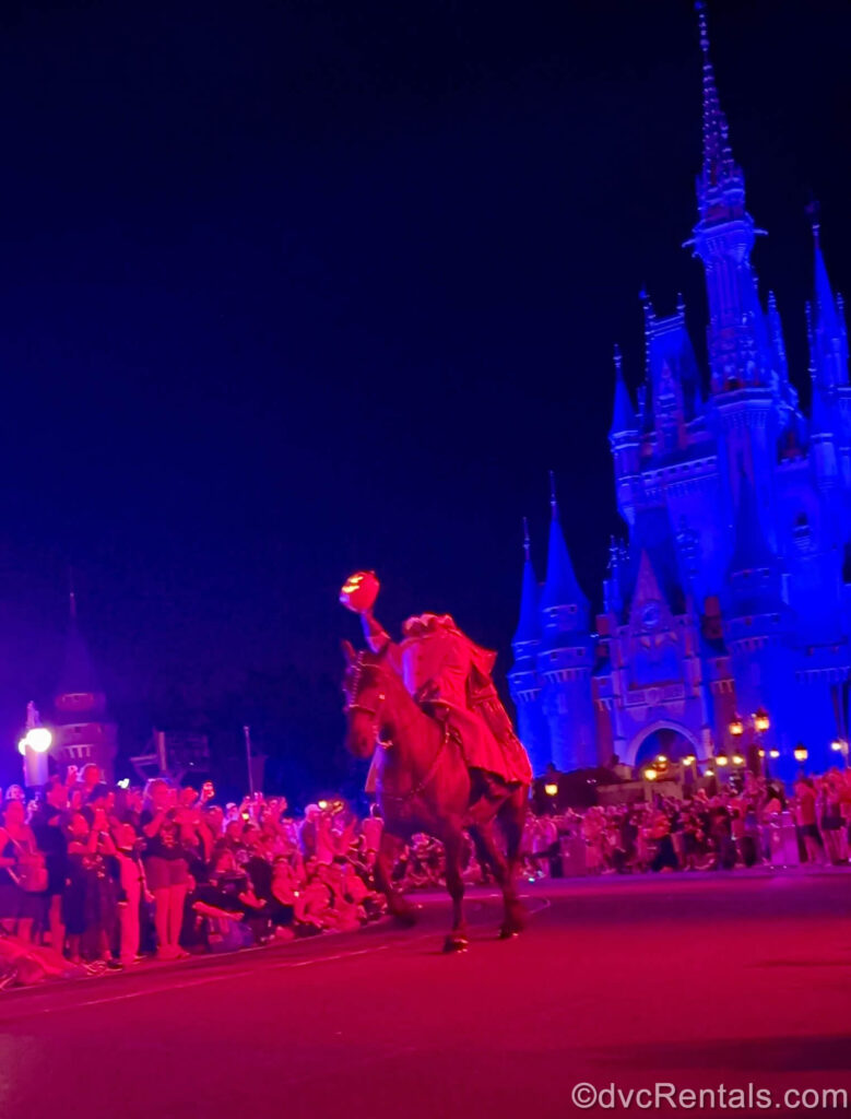 The Headless Horseman holds a glowing jack-o’-lantern as he rides down the parade route at Magic Kingdom at night. Cinderella Castle is lit up blue in the background, while red lighting effects light up the crowds along the sides of the parade pathway.