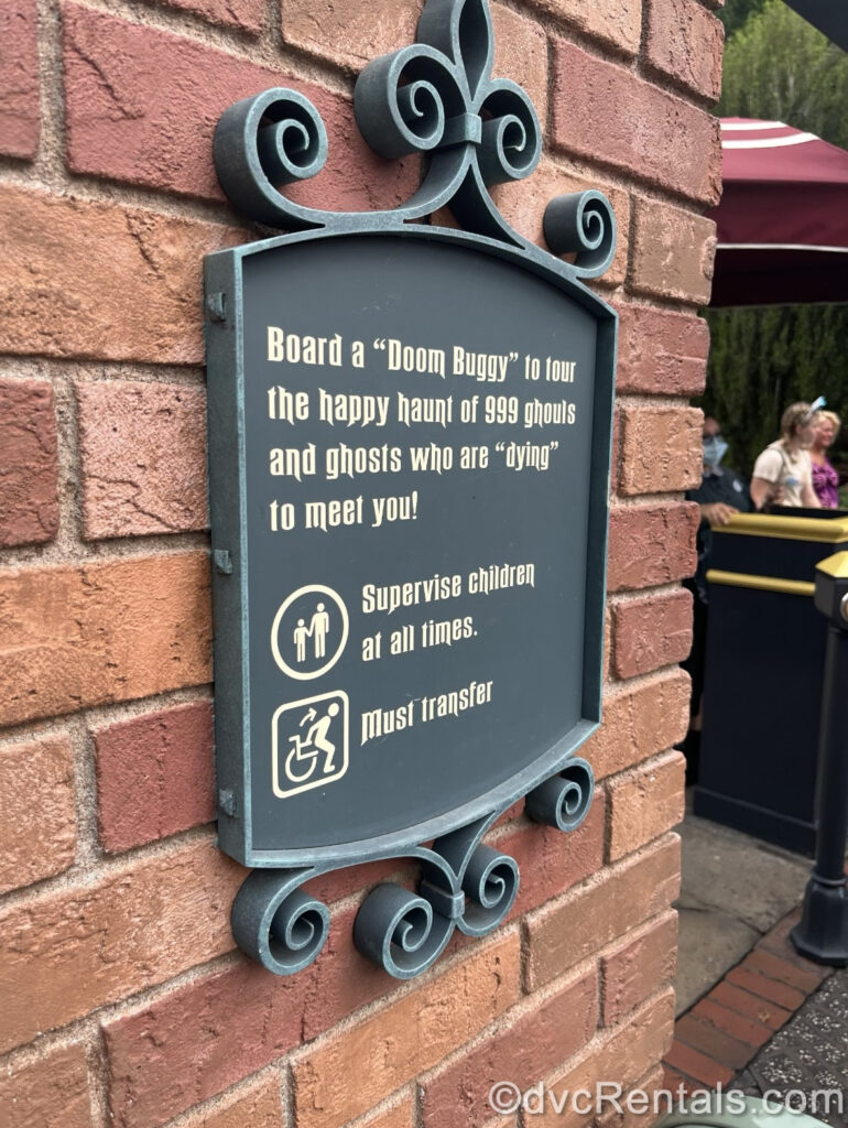 A grey metal sign reading “Board a ‘Doom Buggy’ to tour the happy haunt of 999 ghouls and ghosts who are ‘dying’ to meet you!” in white gothic-looking script is posted on a reddish-brown brick wall in the Haunted Mansion ride queue. The sign also states “supervise children at all times” and advises that those in wheelchairs must transfer to a ride vehicle.