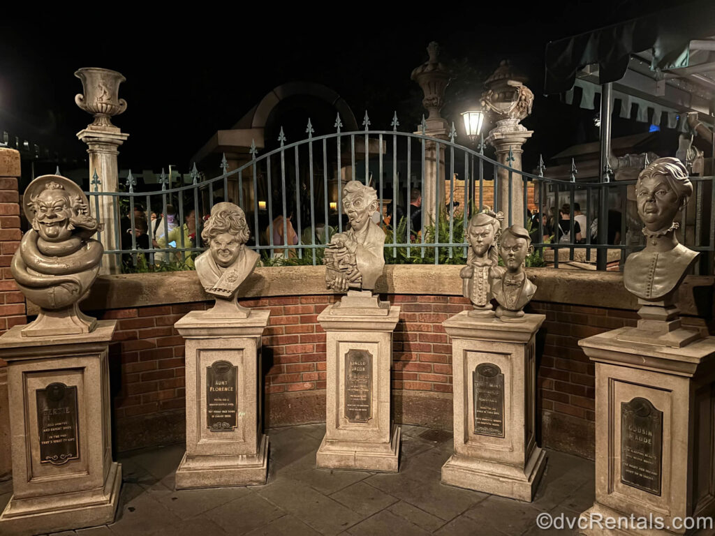 Five marble and stone tomb busts in the Haunted Mansion queue. The busts feature the creepy likenesses of various men, women, and children who were once occupants of the mansion. They are arranged in a semi-circle in front of a reddish-brown brick partition wall and black wrought iron fencing.